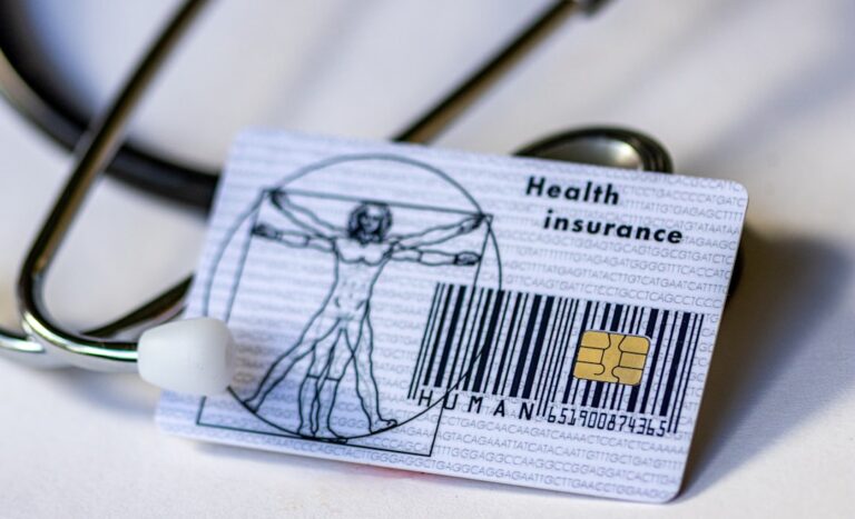 A person sits at a kitchen table reading a denial letter beside a stack of insurance documents and a laptop.